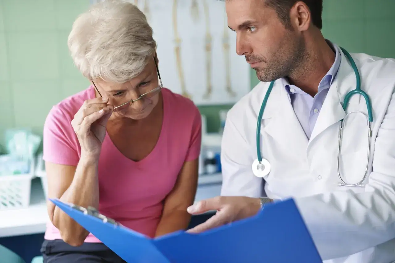 Medico spiega a paziente anziana opzioni per la cura cancro, mostrando documenti su cartella blu.