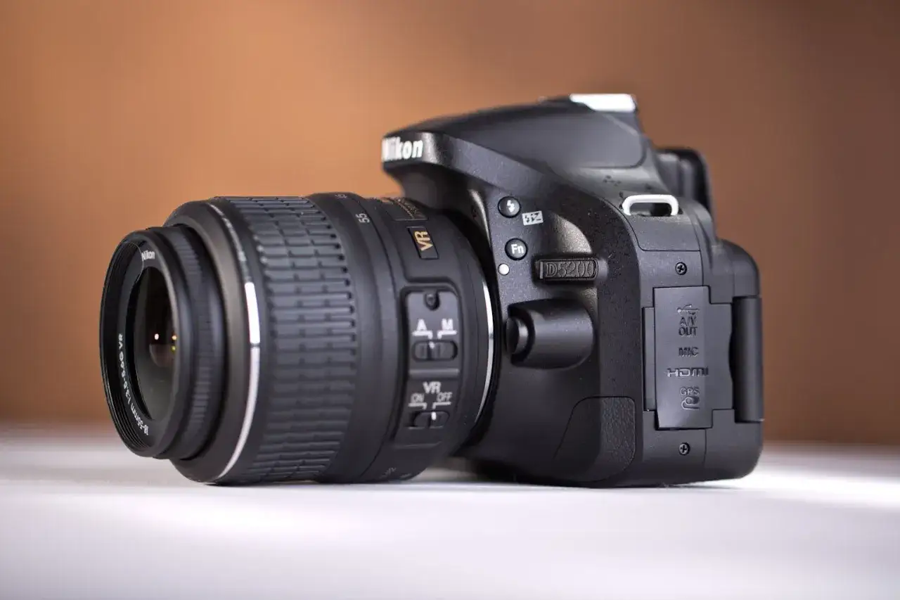 Czarny aparat Nikon D5200 z obiektywem 18-55mm VR. Widoczne przyciski sterujące i złącza.