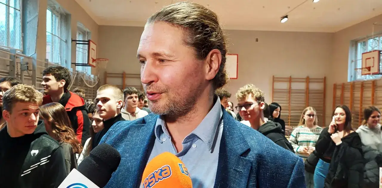 Mężczyzna w garniturze udziela wywiadu, otoczony młodzieżą. Dyskutuje o tym, ile zarabia aktor, w sali gimnastycznej.
