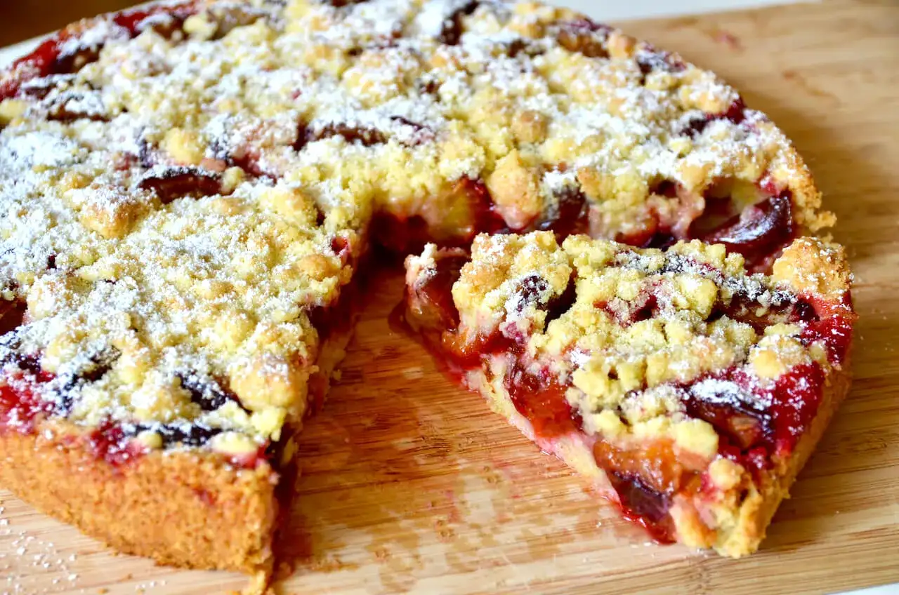 Saftiger Zwetschgenkuchen mit Hefeteig aus der Springform, bestreut mit Puderzucker und Streuseln. Ein Stück ist bereits herausgeschnitten.