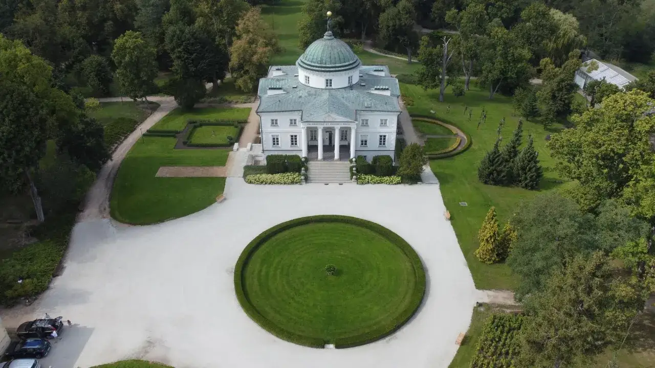 Pałac na Wyspie w Łazienkach Królewskich, otoczony parkiem. W oddali widać szklarnię, a na podjeździe samochody. To miejsce, gdzie historia spotyka się z pięknem, jak w legendach o złotnikach kujawskich.