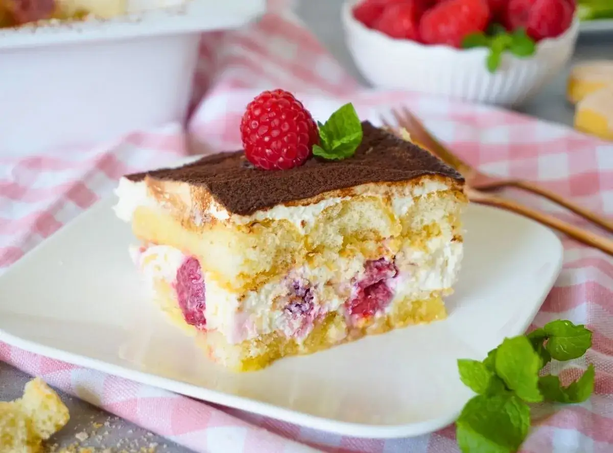 Ein Stück Himbeer Tiramisu, garniert mit einer frischen Himbeere und Minze, auf einem weißen Teller.