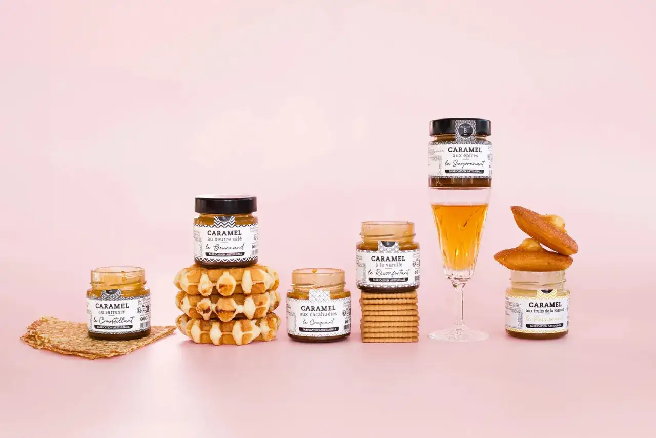 Des pots de caramel artisanal, dont un caramel au beurre sal&eacute;, c&ocirc;toient gaufres, biscuits et madeleines sur fond rose.