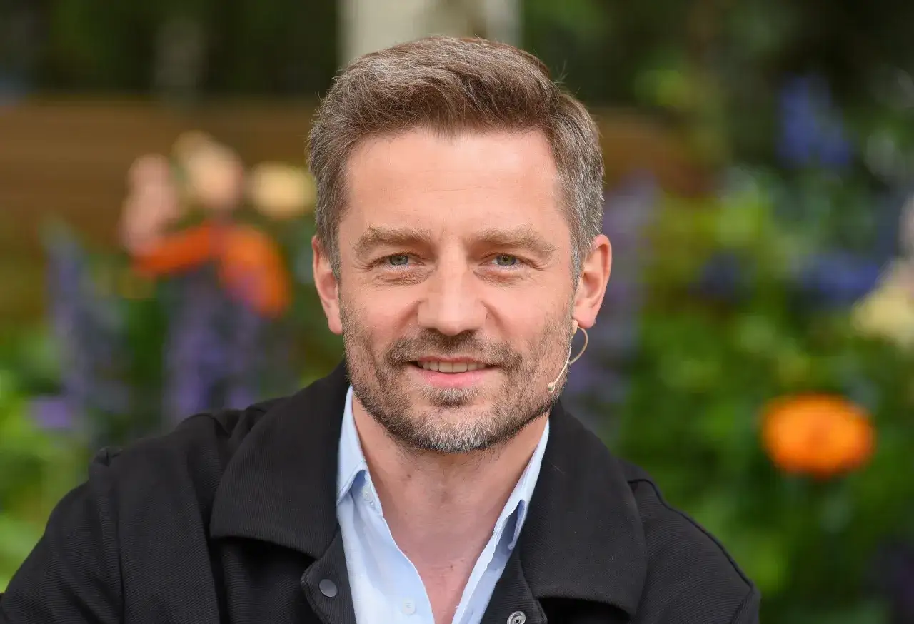 Marcin Rogacewicz, z uśmiechem na twarzy, w otoczeniu zieleni i kwiatów. Myśli o swoich dzieciach.