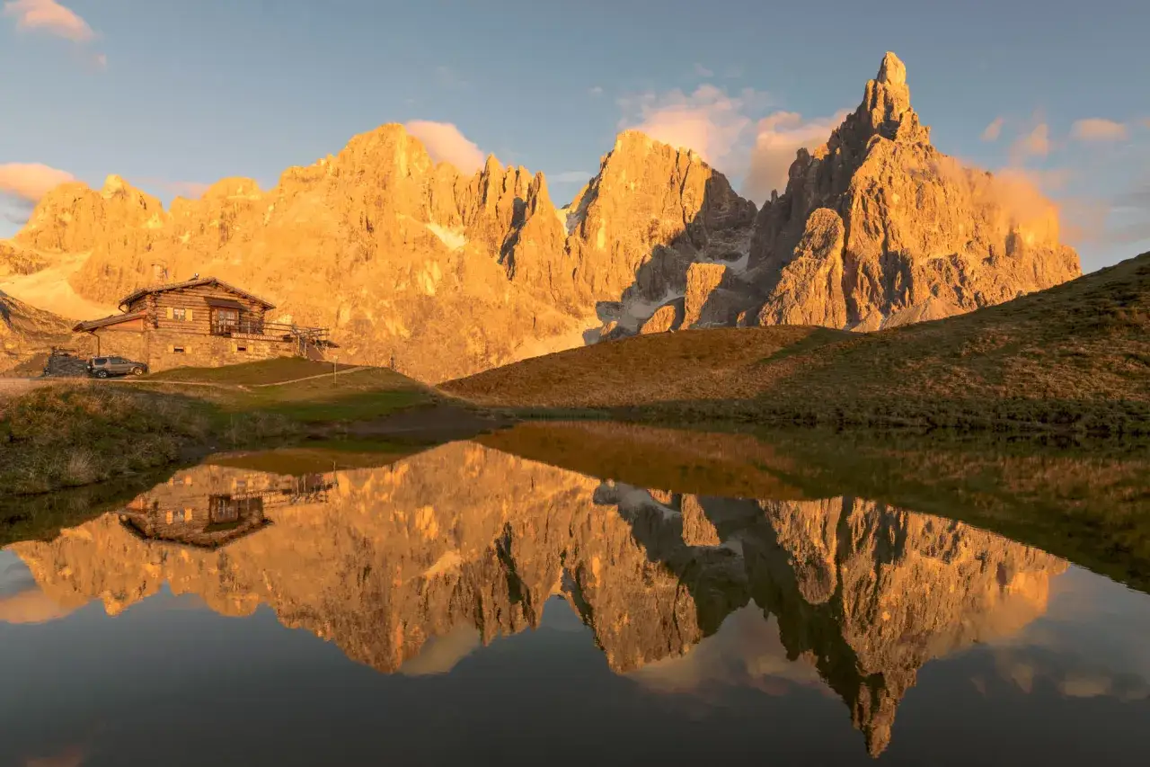 Riflesso perfetto nel lago di un rifugio alpino e del gruppo montuoso delle Dolomiti illuminato dal sole al tramonto.