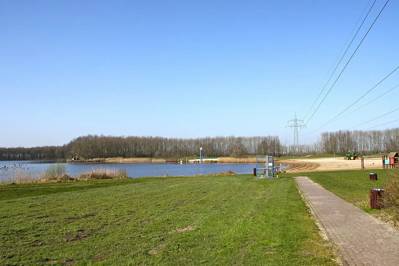 Gr&uuml;ne Wiesen und ein ruhiger Badesee Grotegaste unter blauem Himmel. Ein Traktor ist am Strand zu sehen.