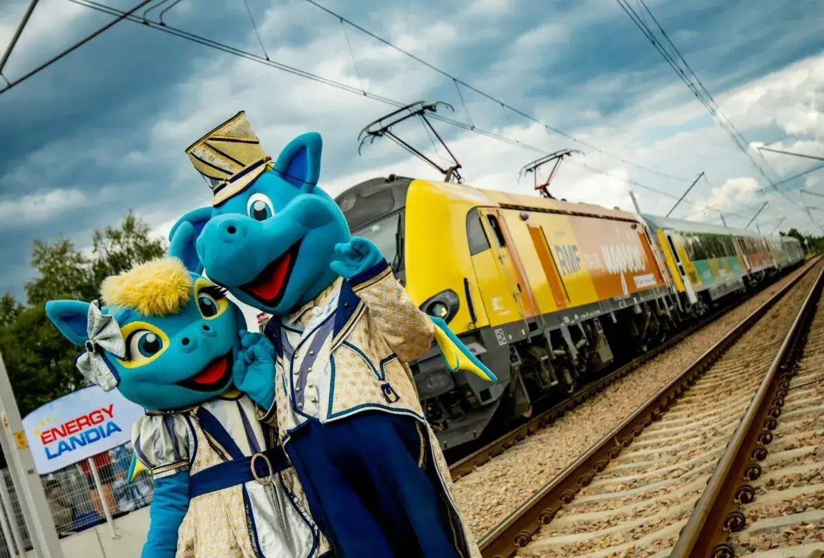 Dwa niebieskie smoki w strojach z epoki pozdrawiają przy pociągu RMF FM. W tle znak Energylandia.
