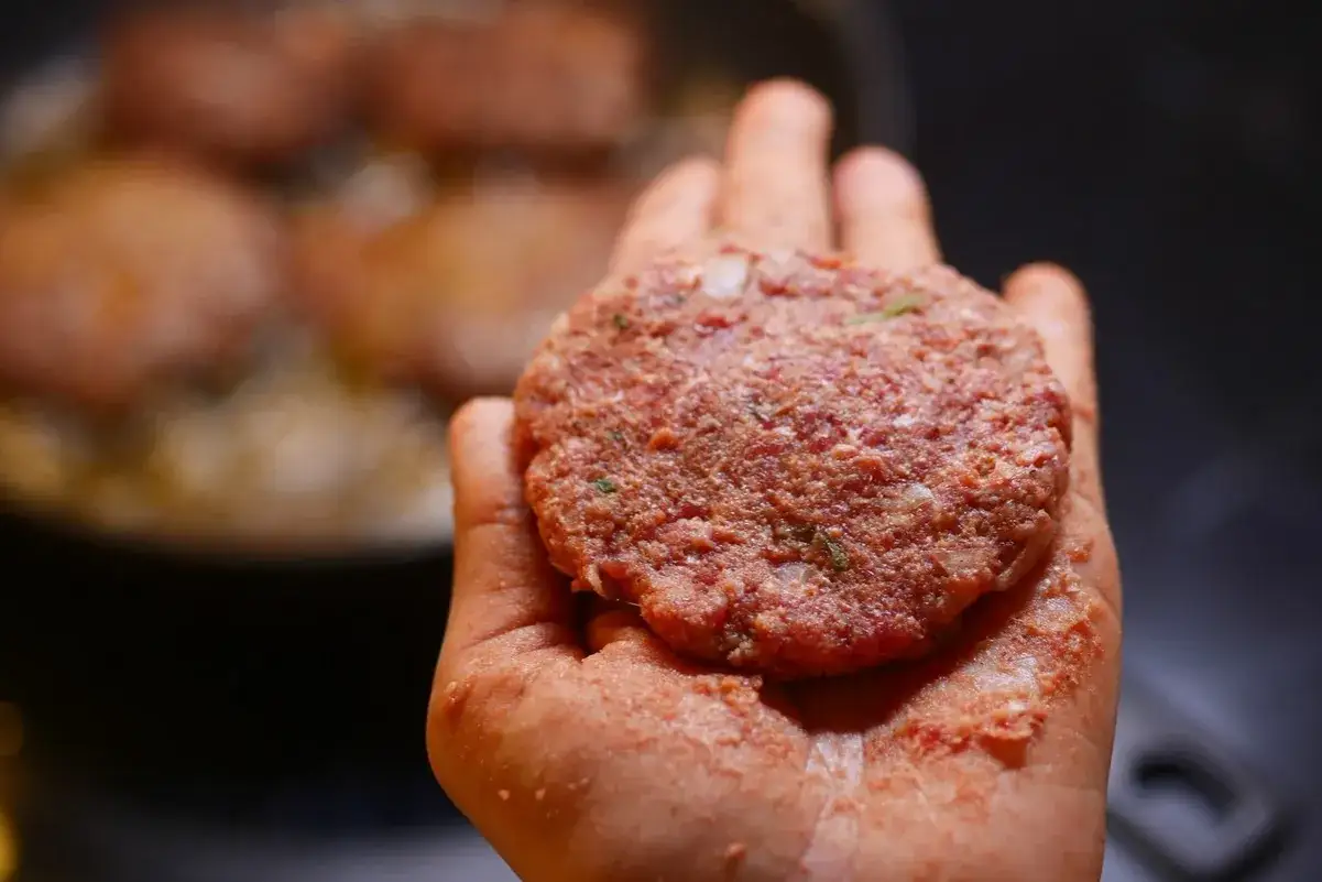 Dłoń trzymająca surowego kotleta do burgera. W tle widać patelnię z innymi kotletami, gotowymi do dalszego etapu, jak składac burgera.