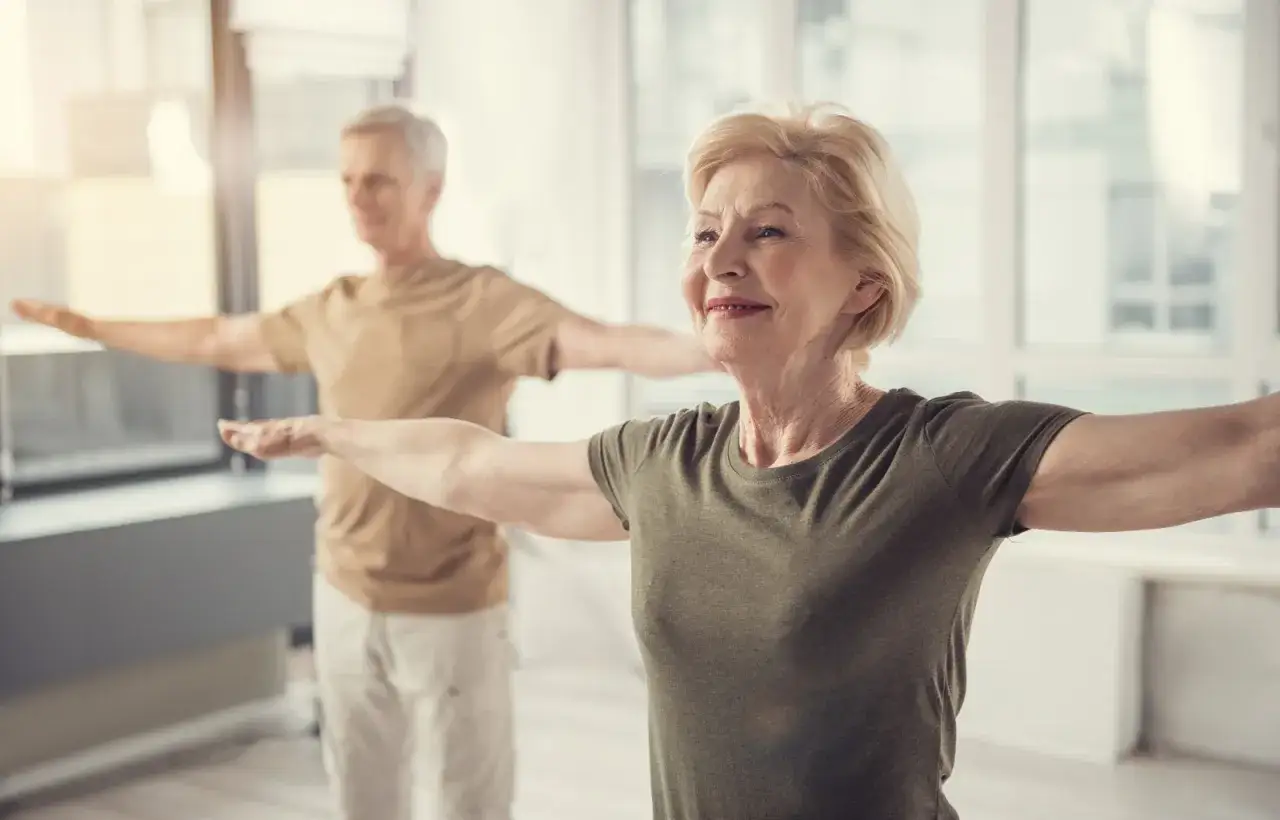 Une femme sportive d'environ 50 ans, souriante, pratique le yoga avec un homme en arrière-plan.