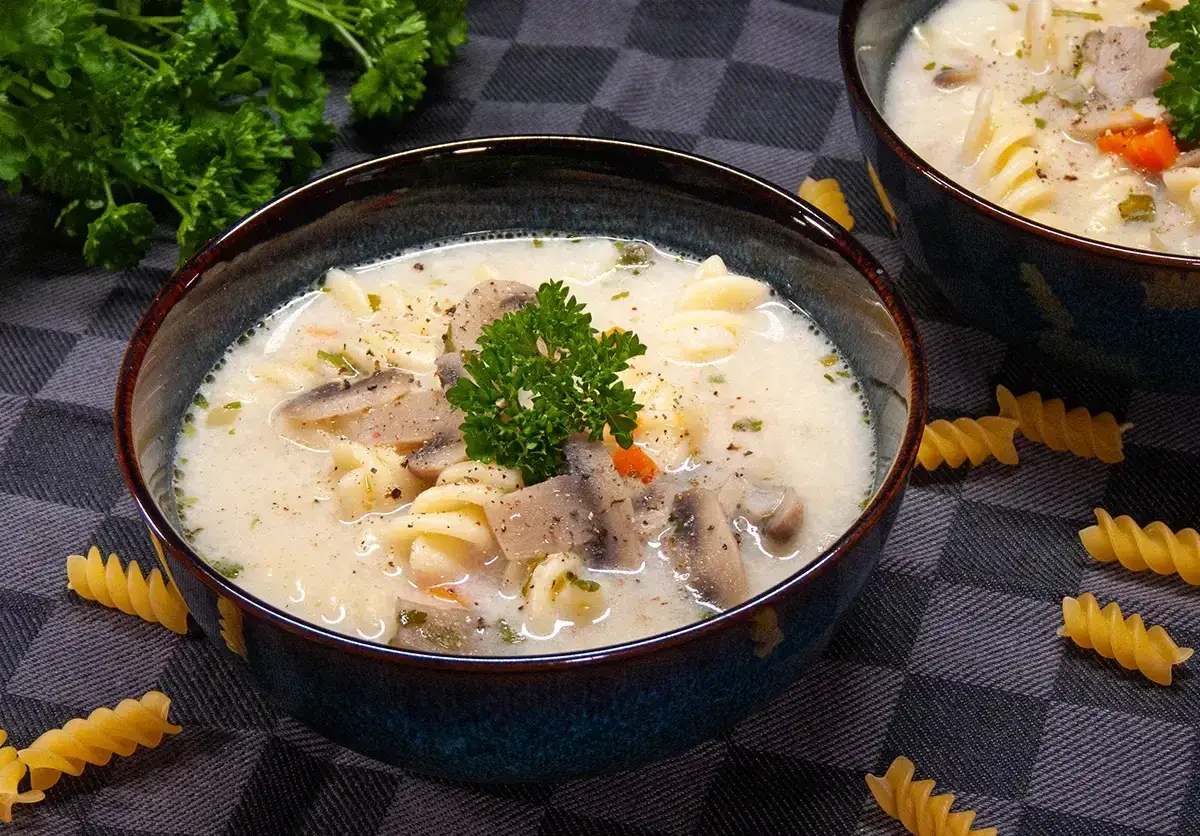 Kremowa zupa z makaronem, grzybami i natką pietruszki. Na stole leżą suche fusilli.