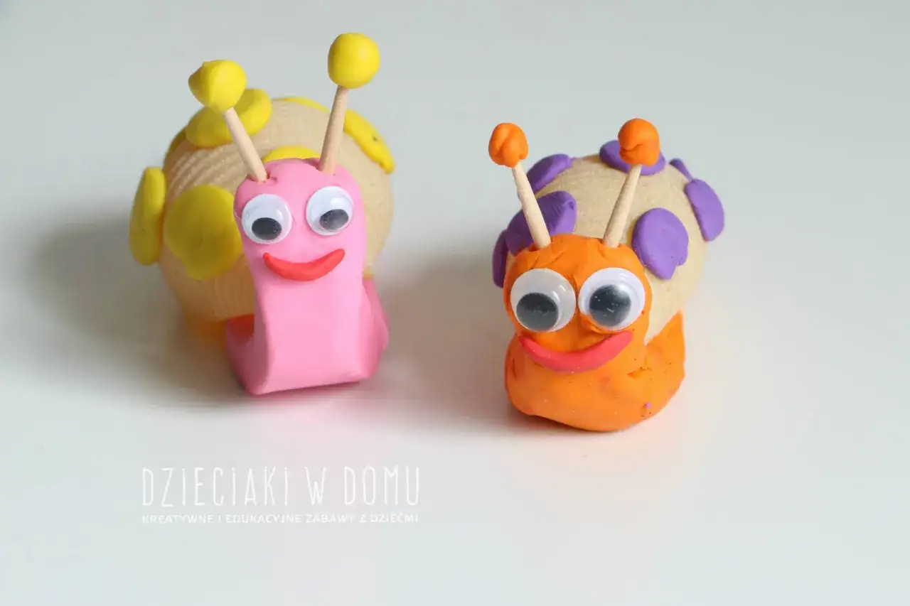Dwa uśmiechnięte ślimaki z plasteliny, jeden różowy, drugi pomarańczowy, z wielkimi oczami i kolorowymi muszlami.