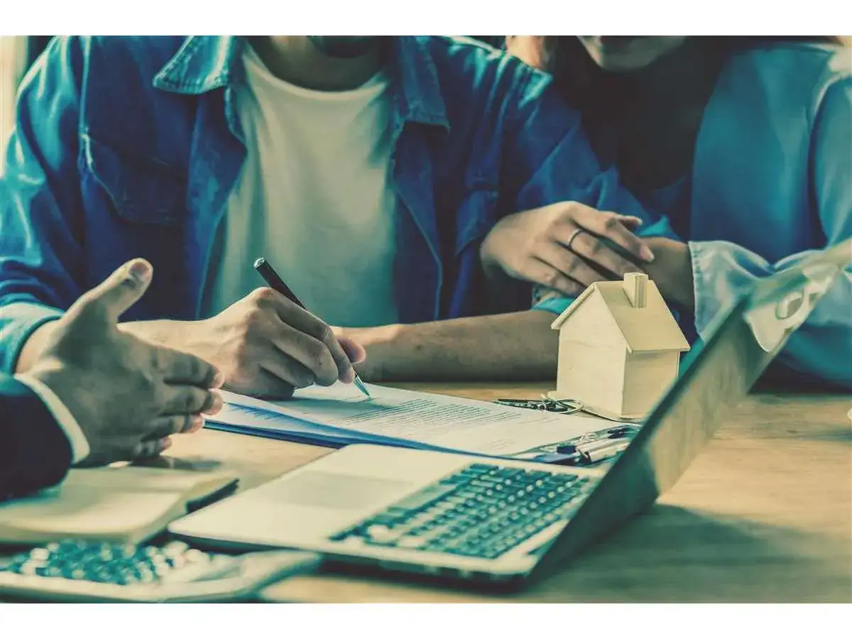 Coppia firma un mutuo con un consulente, con una casa di legno e un laptop. Si discute dopo quanto si può togliere il garante da un mutuo.