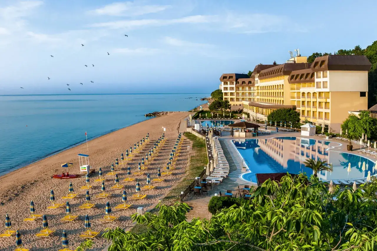 Słoneczne **złote piaski Bułgaria** z rzędami leżaków i parasoli, hotel z basenem i widokiem na morze.