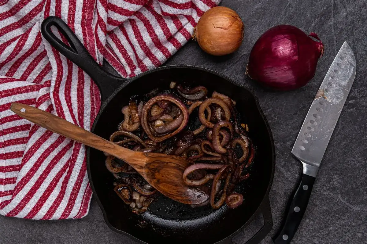 Cipolla rossa in agrodolce, caramellata in padella con un cucchiaio di legno. Accanto, cipolle intere e un coltello.