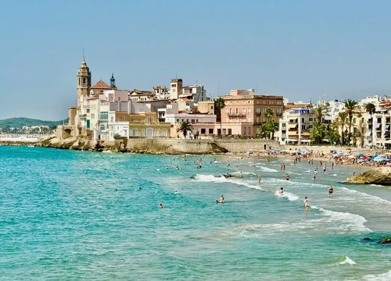 En livlig strand i Sitges, Spanien, med turkost vatten och en stadsk&auml;rna i bakgrunden. M&auml;nniskor njuter av solen och havet.