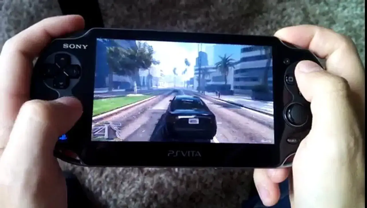 Ręce trzymają PlayStation Vita, grając w GTA. Widok z perspektywy kierowcy czarnego samochodu jadącego ulicą miasta.
