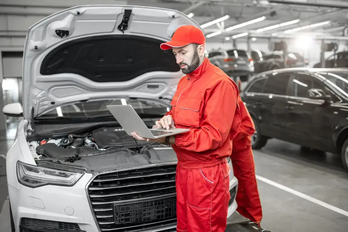 Mechanik w czerwonym kombinezonie sprawdza komputerem diagnostycznym samochód podczas naprawy w ASO.