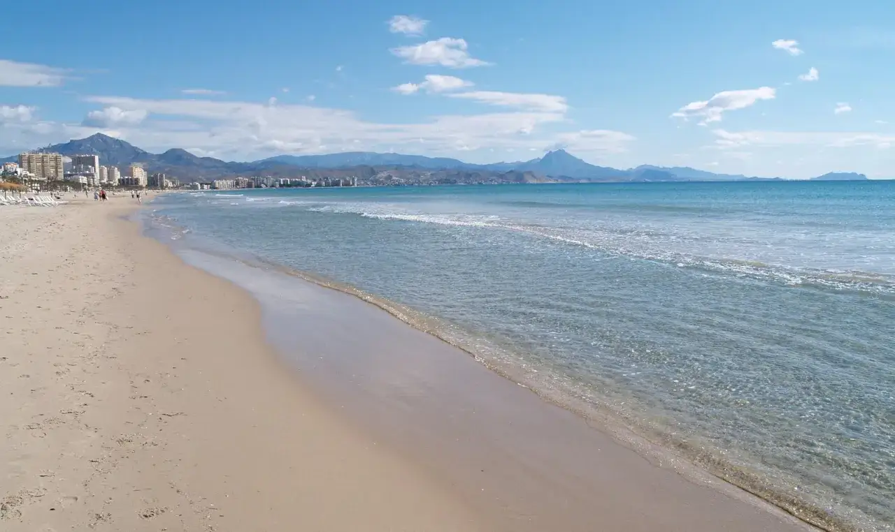 Arena dorada y mar azul en la playa Muchavista, con monta&ntilde;as y edificios al fondo bajo un cielo despejado.