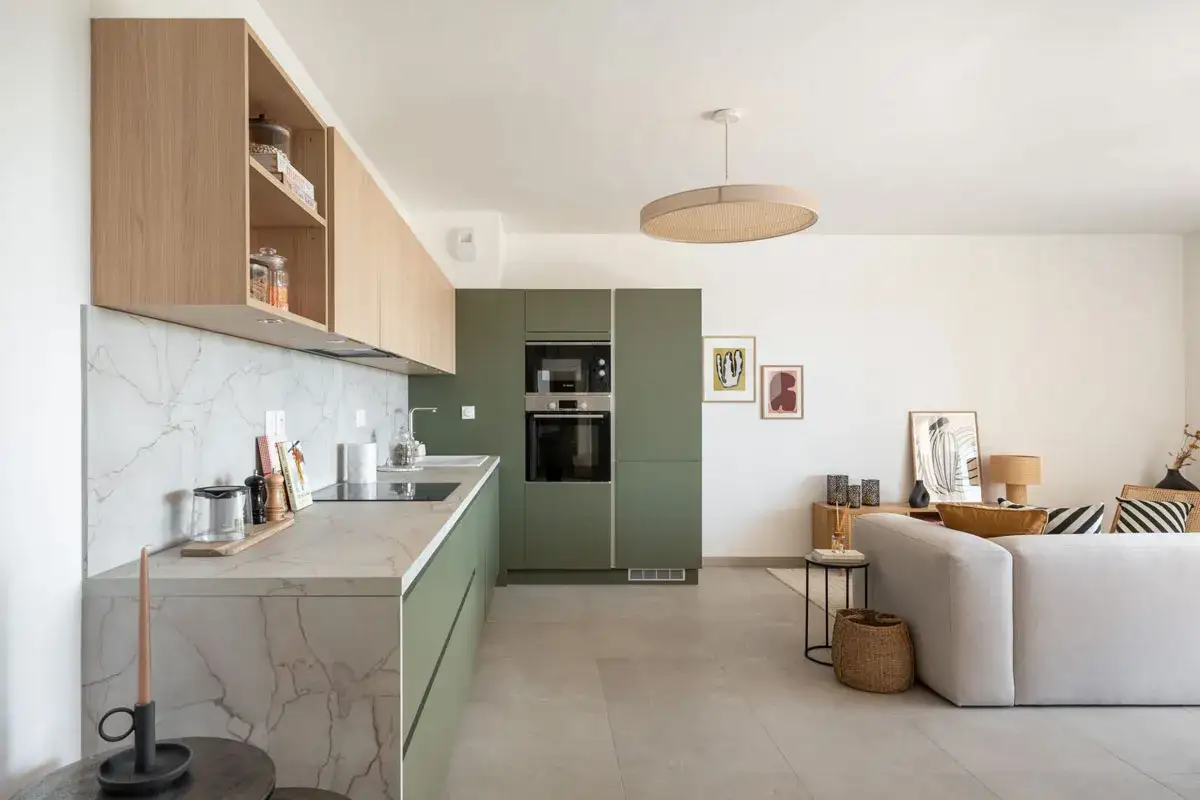 Cuisine moderne avec des armoires vert olive, un plan de travail en marbre et des étagères en bois clair. Un salon est visible à droite.