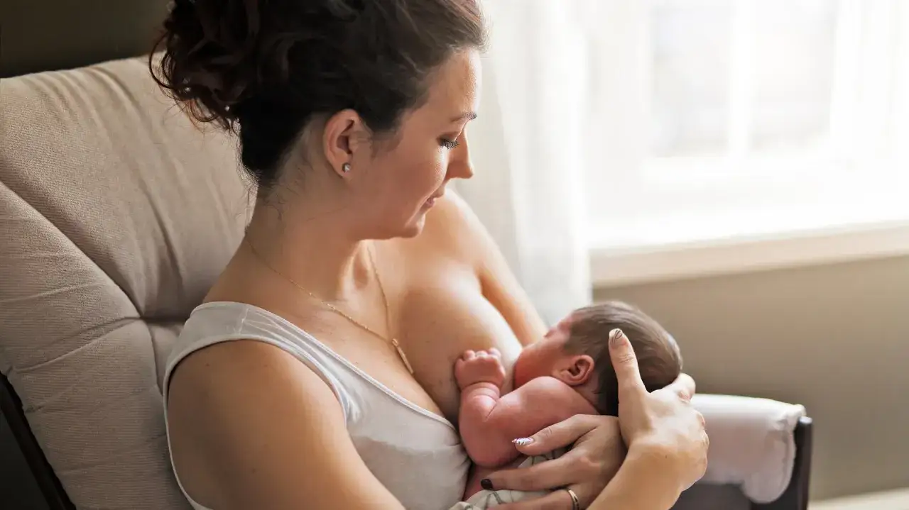Kobieta karmi piersią noworodka, siedząc w fotelu. Delikatny uścisk dłoni, bliskość i miłość. Karmienie piersią w ciąży to piękny moment.