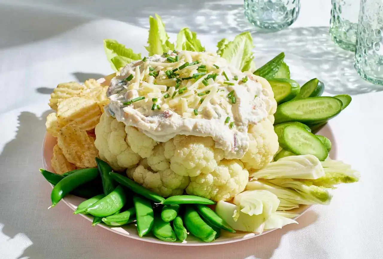 Ein ganzer roher Blumenkohl als Dip-Schüssel, umgeben von Erbsen, Gurken, Chips und Salat. Perfekt, um blumenkohl roh essen zu genießen.