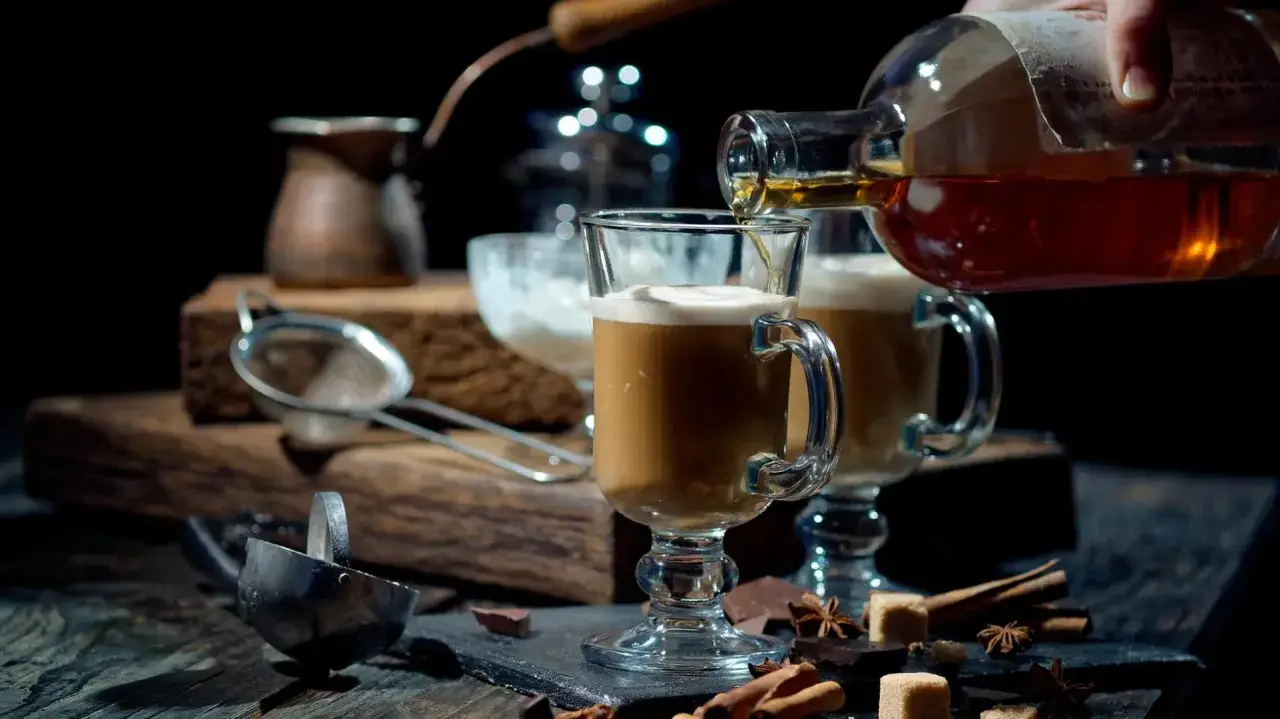 Ein Glas heißer Kaffee mit Sahne, dem gerade ein Schuss Likör zugegeben wird. Dazu Schokolade und Zimt.