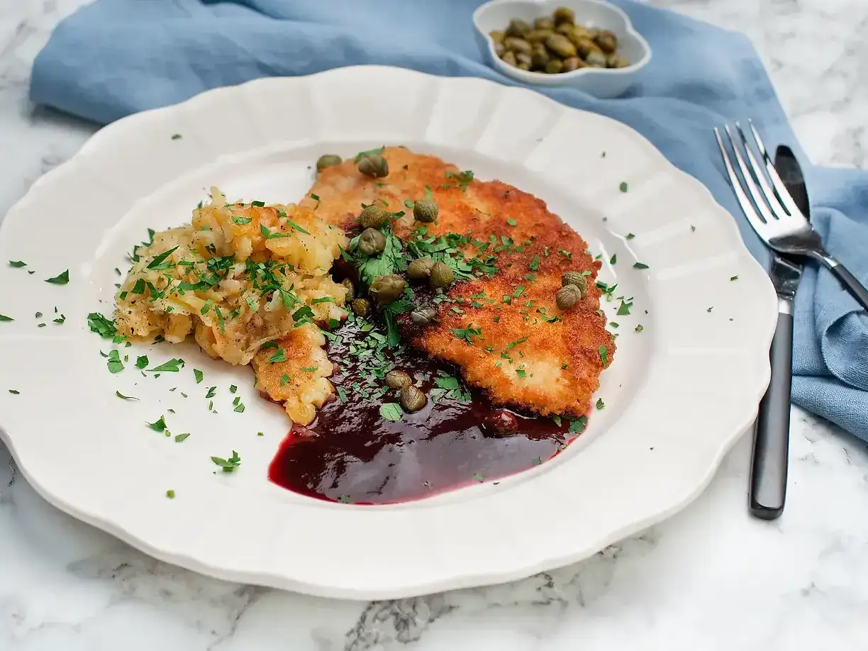 En tallrik med schnitzel, potatis och en mörk sås till schnitzel, garnerad med kapris och persilja.