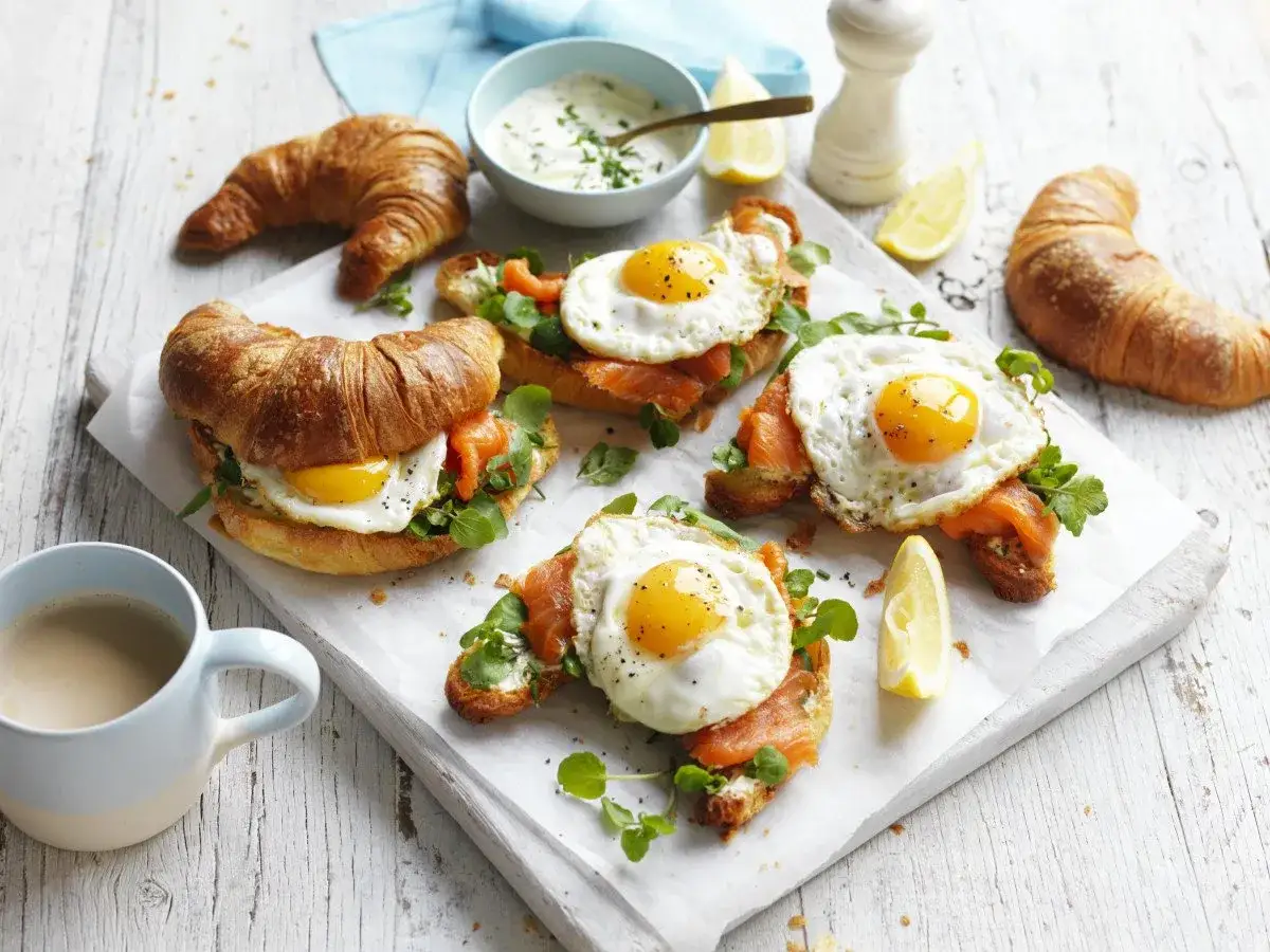 Szybkie śniadanie na ciepło: kruche croissanty z łososiem, jajkiem sadzonym i rukolą. Obok kawa i sos.