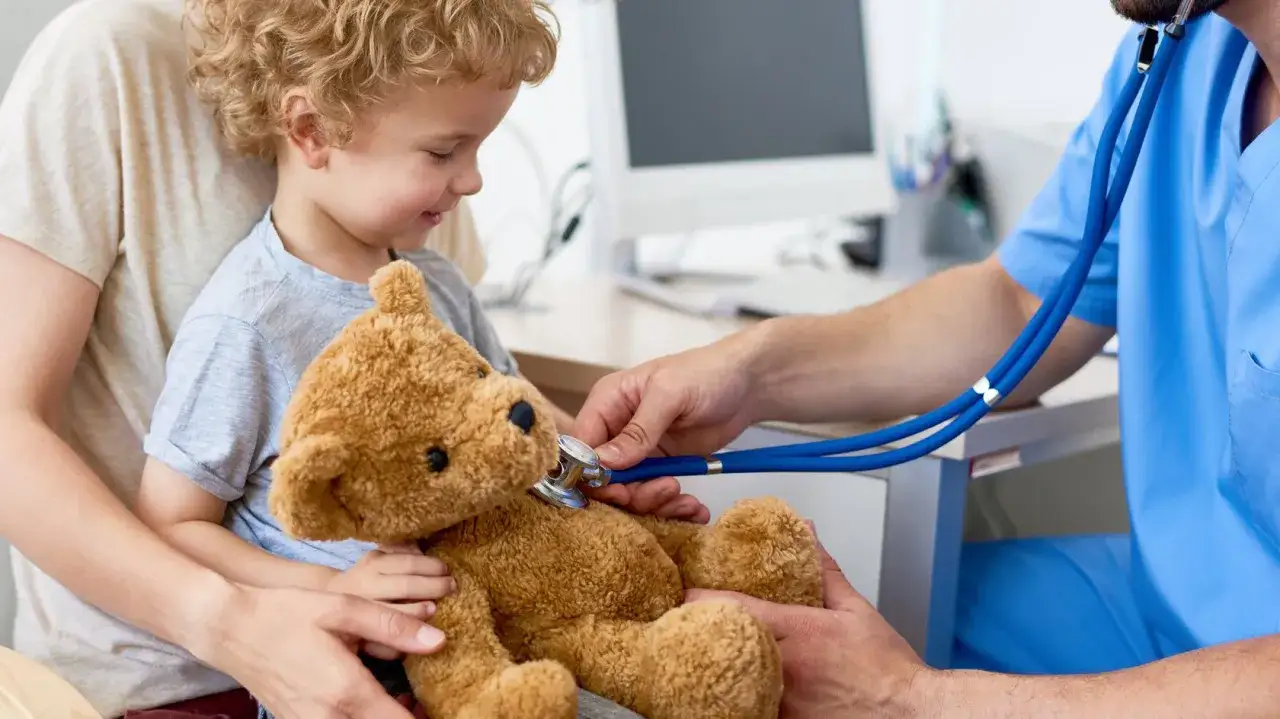 Arzt untersucht Teddyb&auml;r eines Kindes. Die Mutter h&auml;lt ihr Kind, das leicht besorgt wirkt.