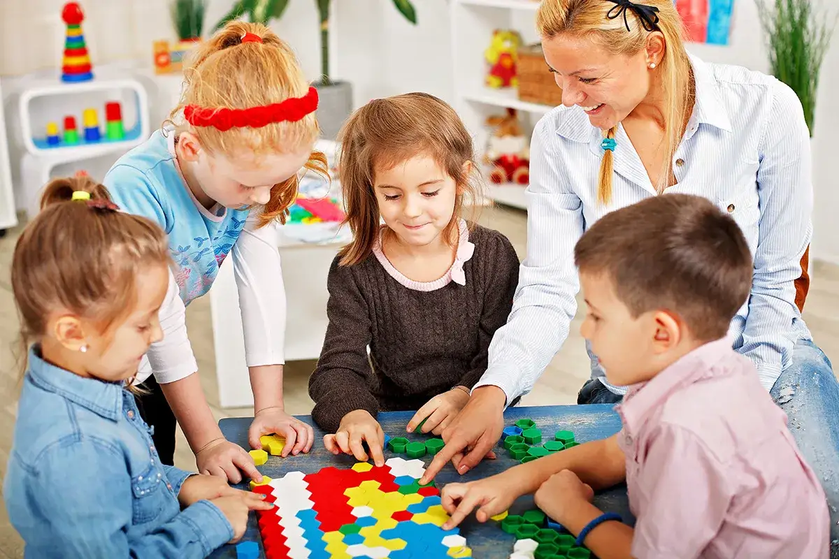 Bambino 5 anni e altri bambini giocano con tessere colorate, guidati da un'insegnante sorridente.