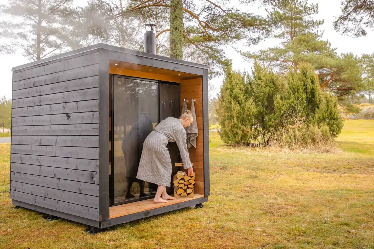 En kvinna i morgonrock laddar ved vid en modern bastu. En bra investering f&ouml;r att veta bygga bastu kostnad.
