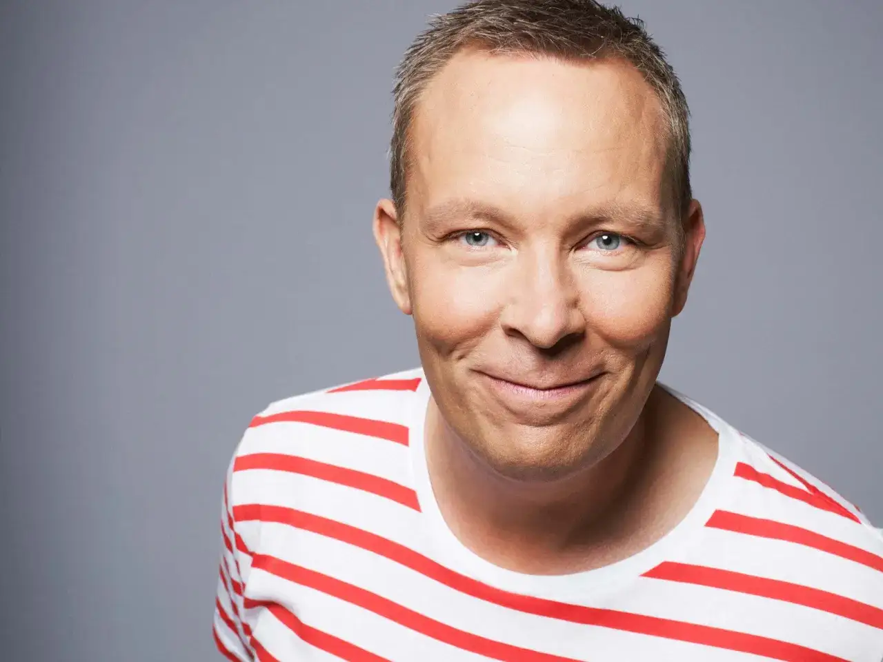 En leende man, Stellan Sundahl, med bl&aring; &ouml;gon och kort, gr&aring;tt h&aring;r, b&auml;r en vit t-shirt med r&ouml;da r&auml;nder.