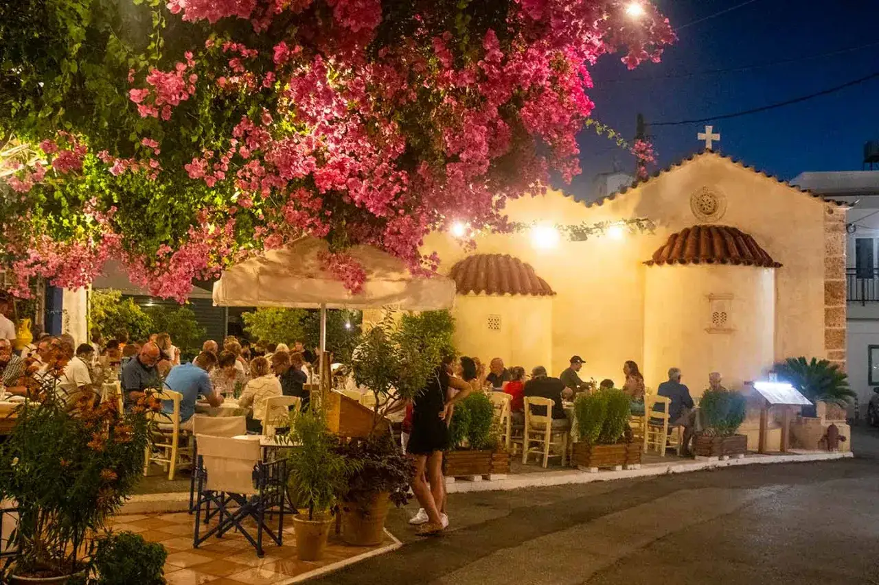 Abendessen unter Bougainvillea in Malia, Kreta. Gäste genießen die Atmosphäre vor einer Kirche.