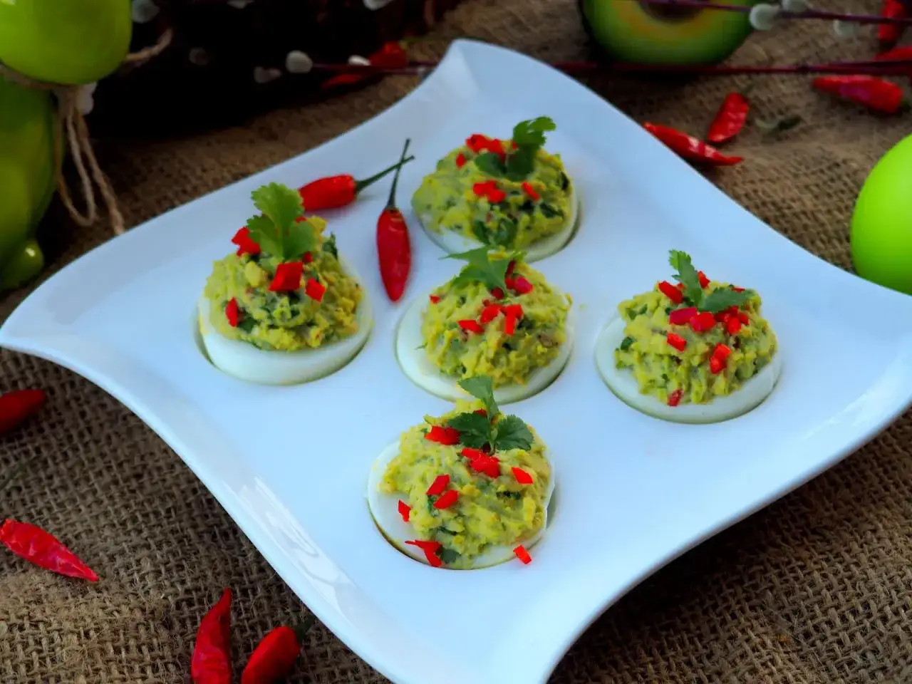 Jajka faszerowane guacamole, ozdobione papryczką chili i kolendrą, prezentują się apetycznie na białej tacy.