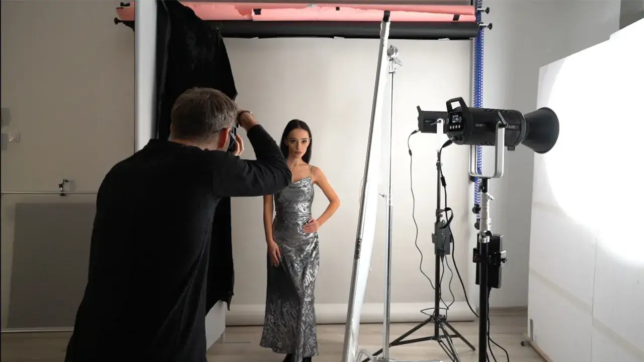 Sesja zdjęciowa: fotograf ustawia lampy w studio, modelka w srebrnej sukni pozuje na tle białego tła.