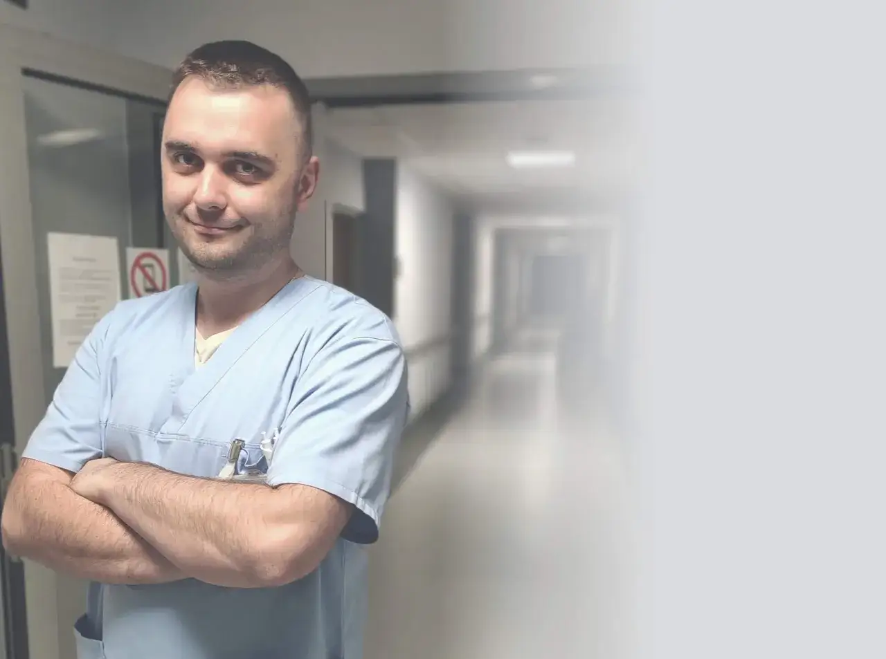 Mężczyzna w niebieskim uniformie medycznym stoi ze skrzyżowanymi rękami na tle szpitalnego korytarza.