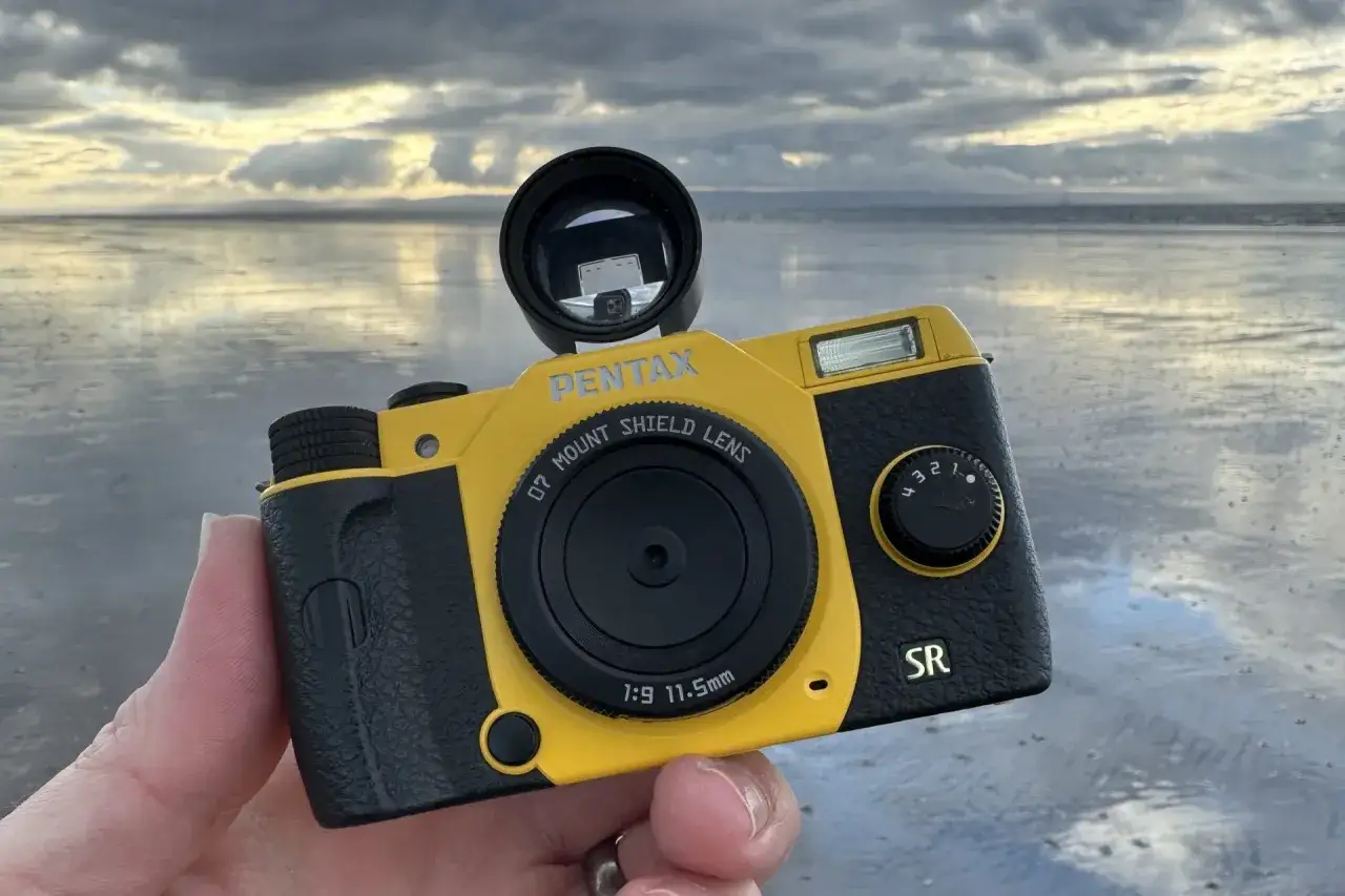 En gul och svart Pentax Q-kamera h&aring;lls upp mot en molnig himmel och en spegelblank strand.