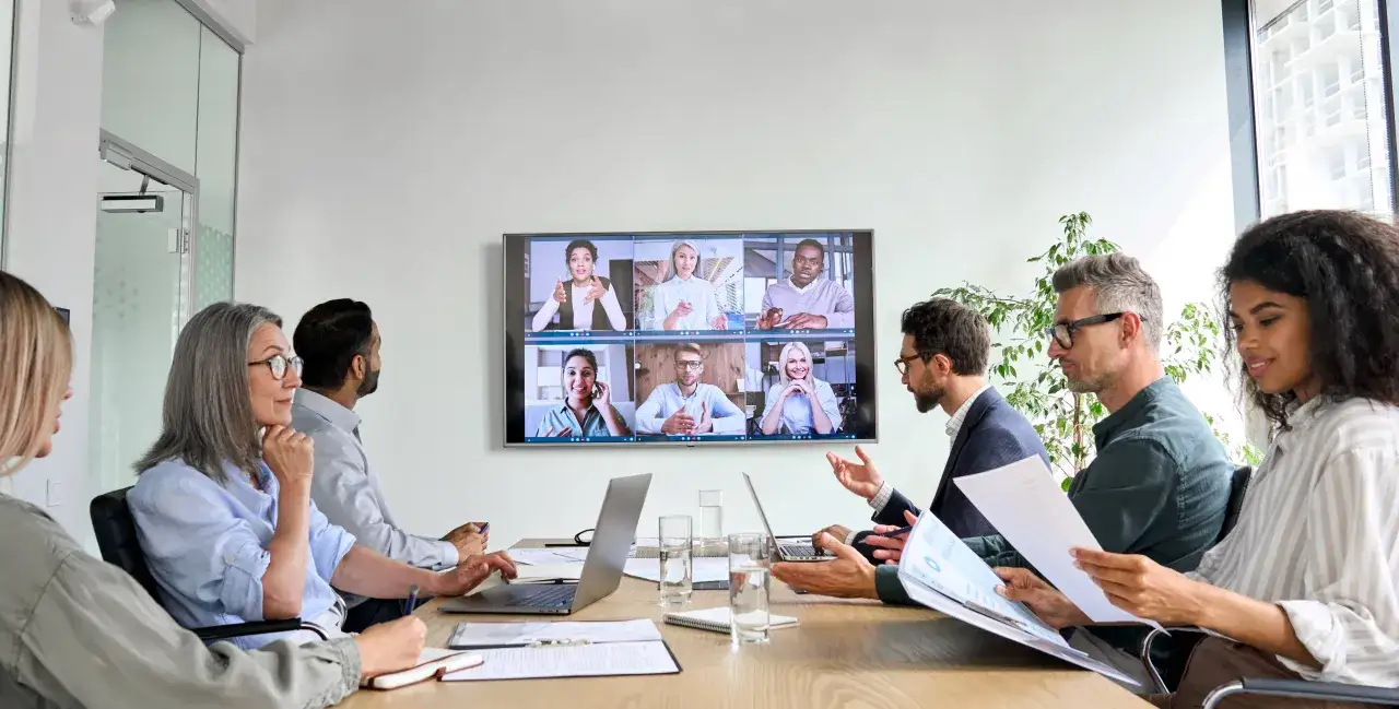 Equipo en una reuni&oacute;n de pie, colaborando en un proyecto con colegas en pantalla.
