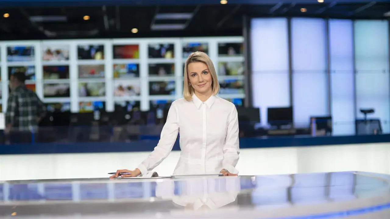 Joanna Dunikowska-Paź, znana z Wikipedii, w białej koszuli stoi przy stole w studiu telewizyjnym.