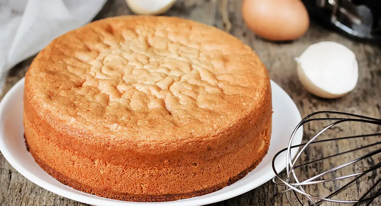 Ein goldbrauner Tortenboden, perfekt für ein Tortenboden Rezept 3 Schichten. Eier und Schneebesen deuten auf die Zubereitung hin.
