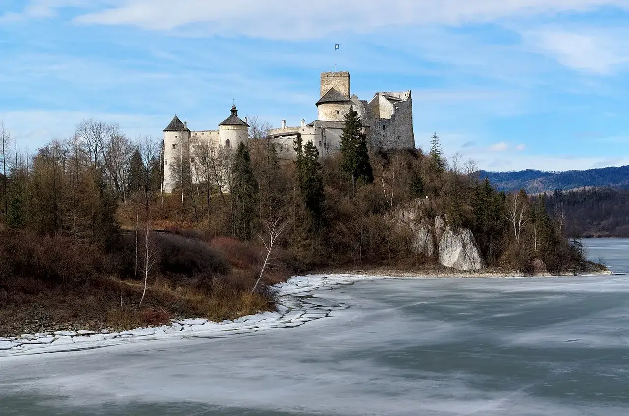 Zamek Dunajec majestatycznie wznosi się na wzgórzu nad zamarzniętym jeziorem, otoczony zimową przyrodą.