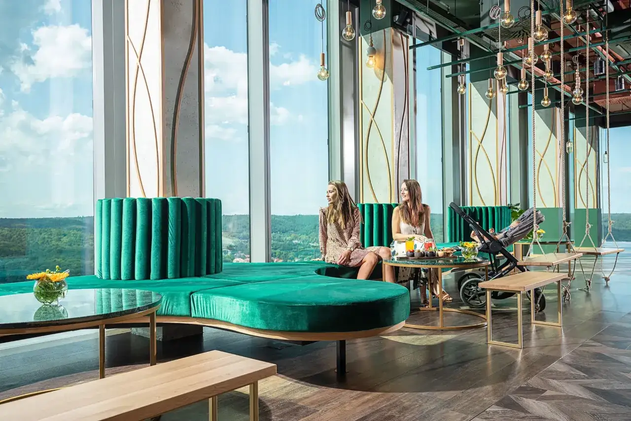 Dwie kobiety relaksują się w nowoczesnym bistro na piętrze, podziwiając widok przez ogromne okna. Wygodne, zielone kanapy i huśtawki tworzą przytulną atmosferę.