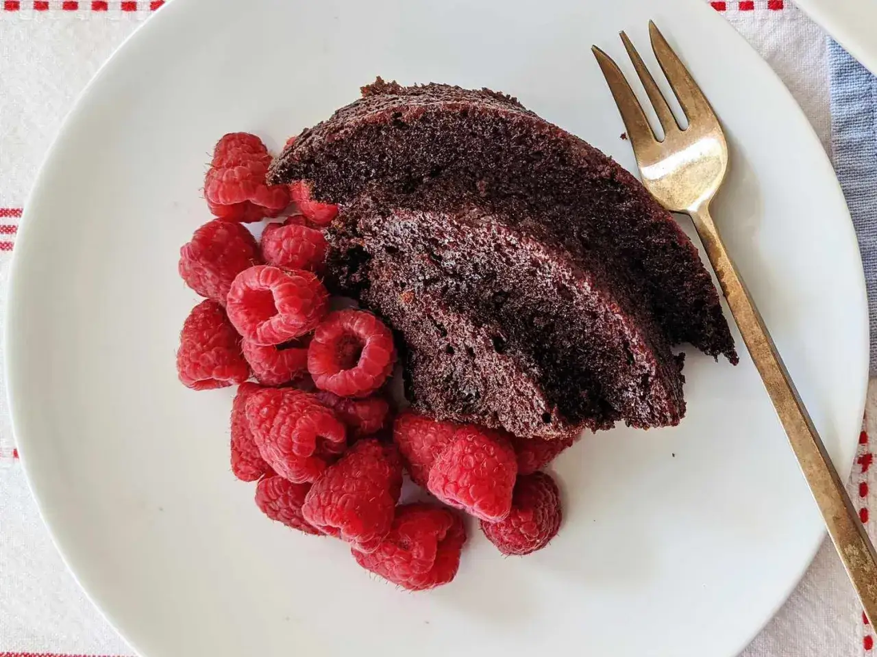 Zwei St&uuml;cke saftiger veganer Schokokuchen mit frischen Himbeeren und einer goldenen Gabel auf einem wei&szlig;en Teller.