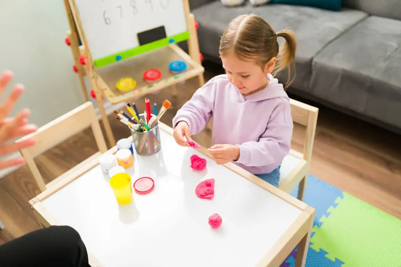 Ni&ntilde;a concentrada jugando con plastilina rosa, explorando sus emociones infantiles. A su lado, pinturas y l&aacute;pices de colores.