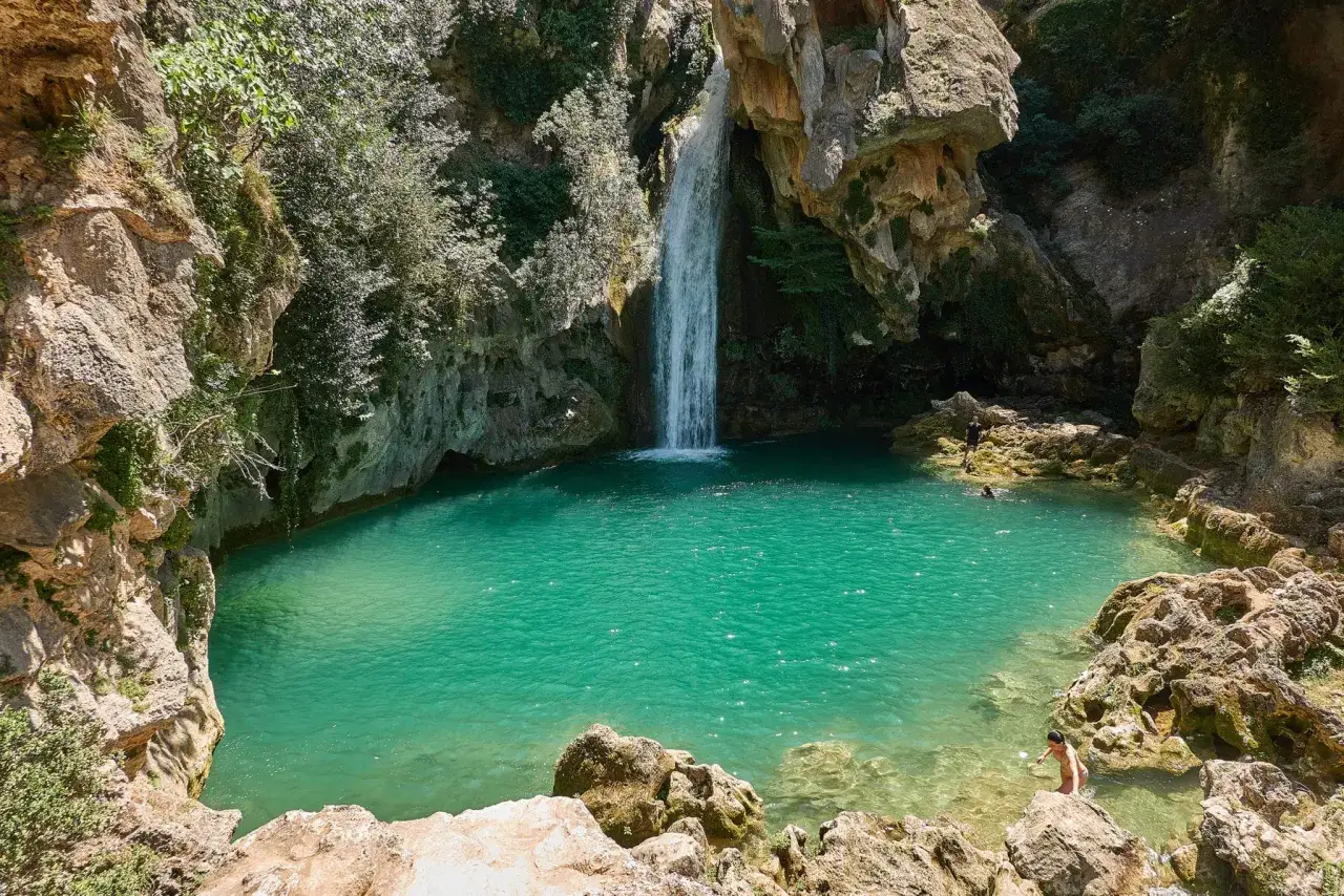 La cascada de la calavera desciende a una poza turquesa rodeada de rocas y vegetación. Personas disfrutan del agua cristalina.