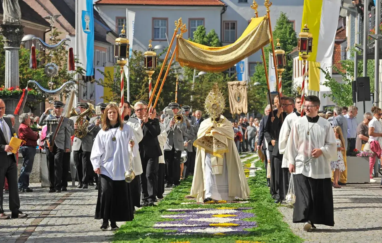Prozession am Fronleichnam Feiertag. Gläubige ziehen über einen Blumenteppich, begleitet von Musikern und Fahnen.