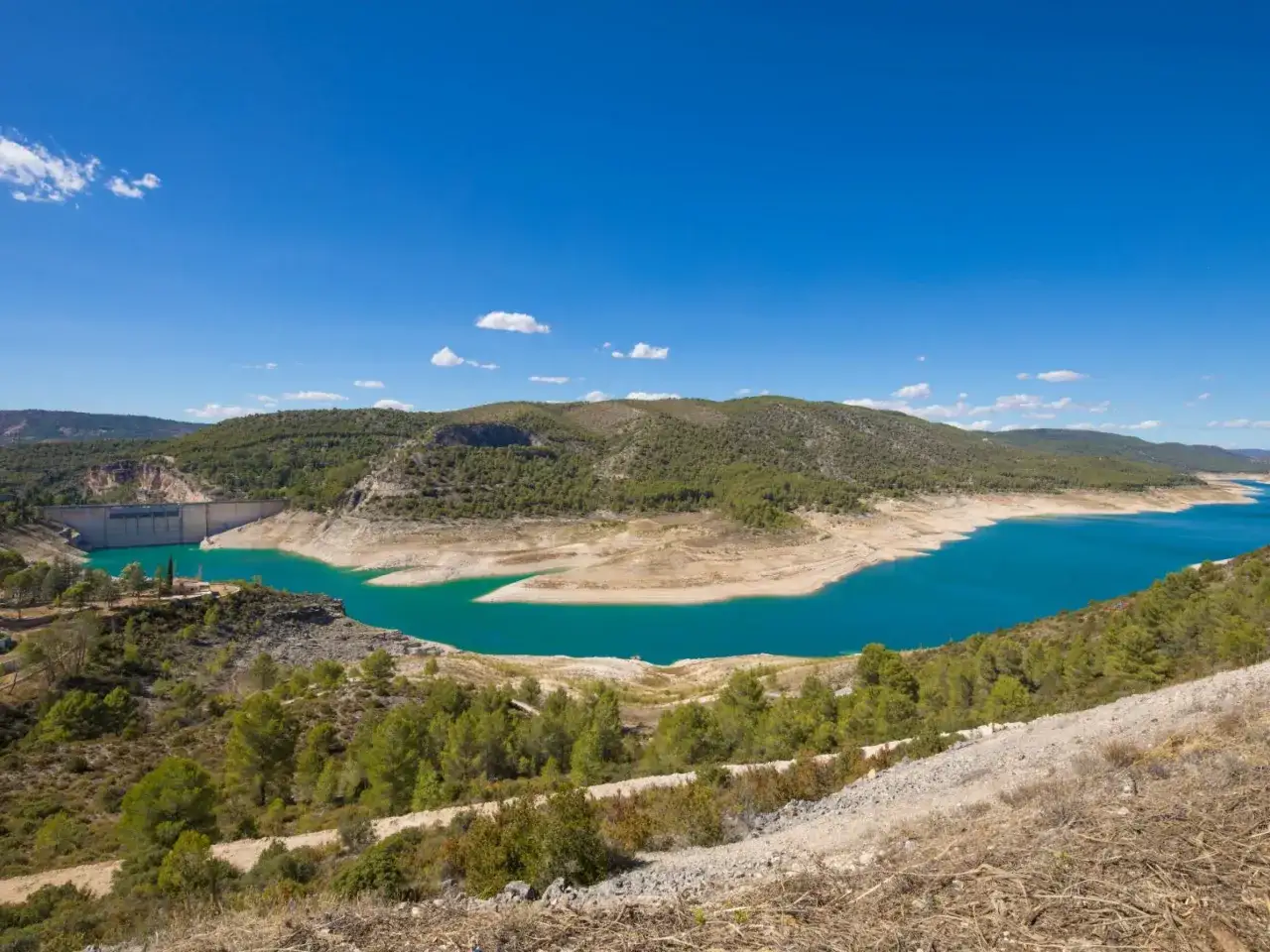El pantano de Saced&oacute;n, con su agua turquesa, se extiende entre colinas verdes bajo un cielo azul.