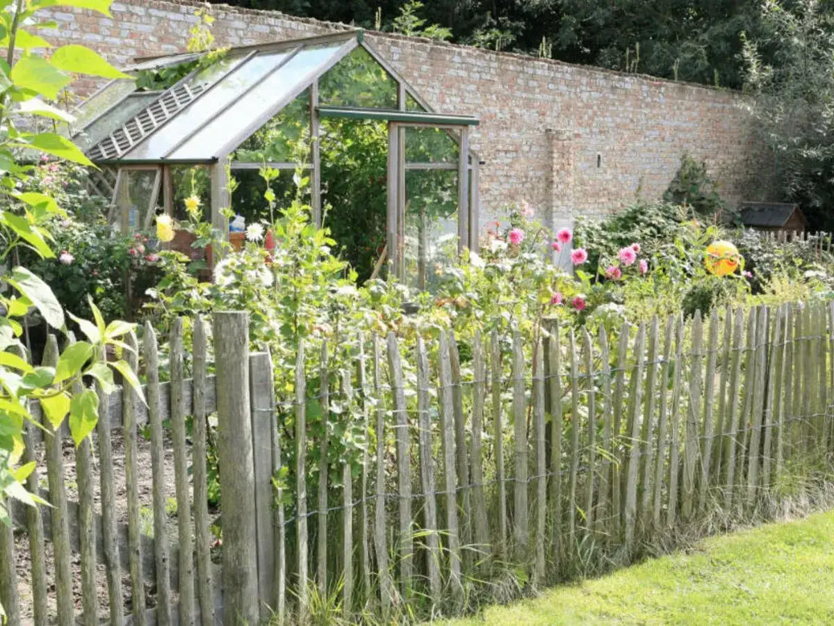 Ein rustikaler staketenzaun befestigen, um einen bl&uuml;henden Garten mit Gew&auml;chshaus und Ziegelmauer zu sch&uuml;tzen.