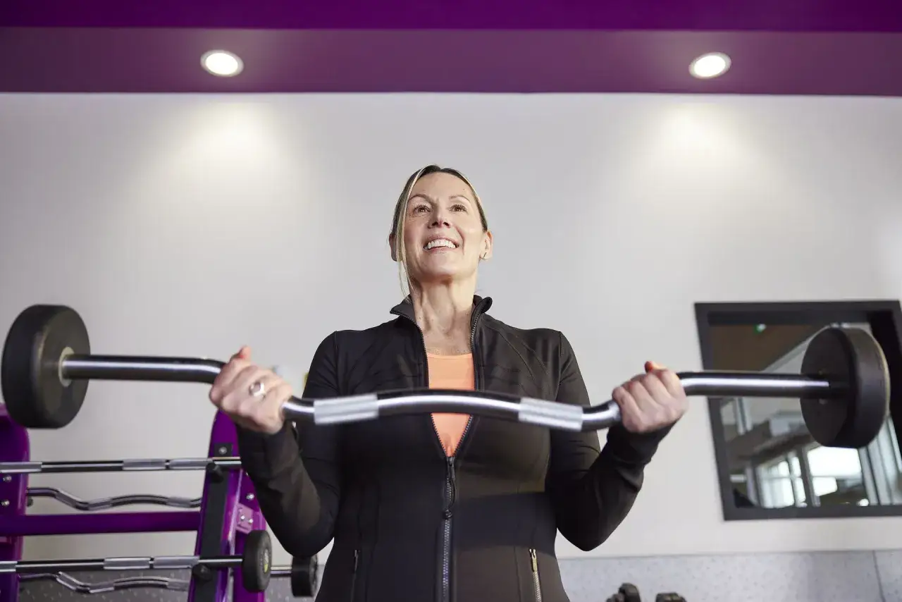Uśmiechnięta kobieta wykonuje pompki na biceps z gryfem. W tle stojaki na ciężary.