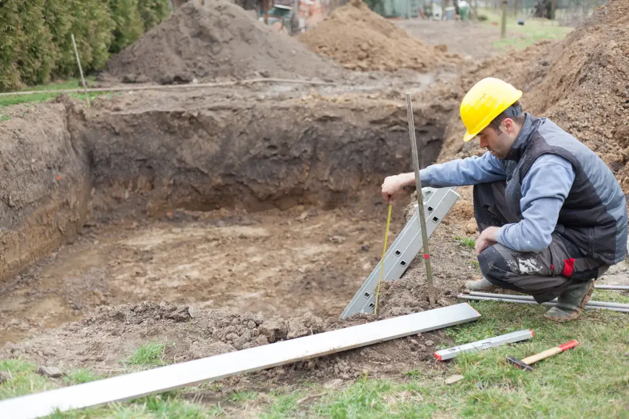 Budowlaniec w żółtym kasku mierzy głębokość wykopu pod fundament. Wokół leżą narzędzia i materiały budowlane.